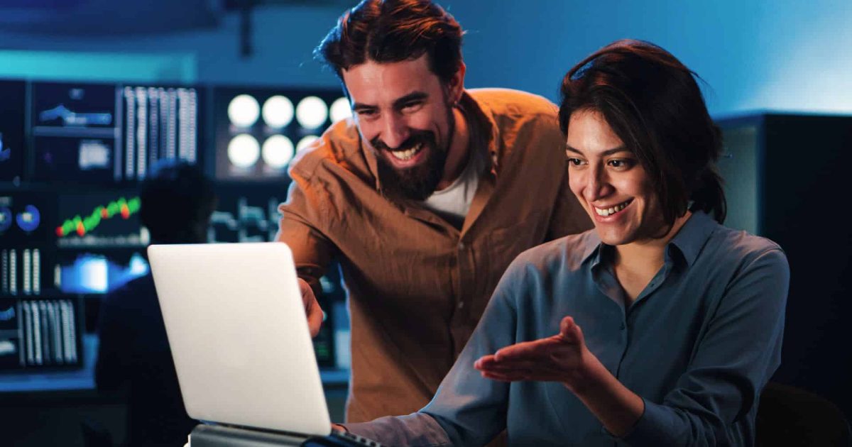 Happy business couple working on a laptop computer in the office. Young trading team people smiling watching data on pc screen. Two partners coworking on finance. High quality photo