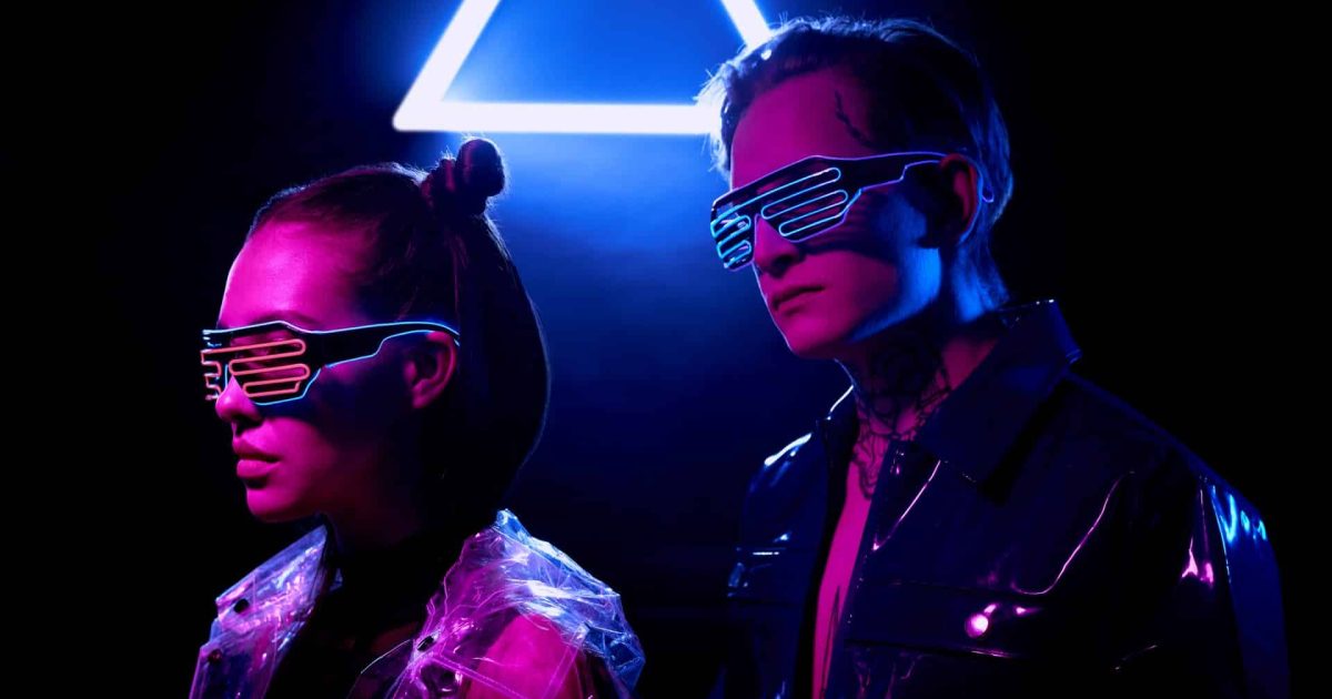 Young cyber couple in vinyl jackets and LED goggles standing in dark room with neon triangle sign