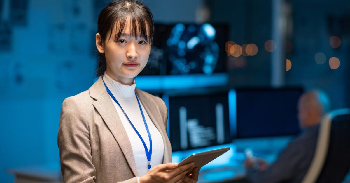 Young serious female programmer or developer in formalwear holding tablet while networking in office environment